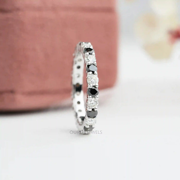 A close-up image of a Round Cut Lab Diamond Eternity Wedding Band featuring white and black lab-grown diamonds ring. It displayed against a soft pink background.