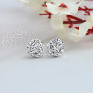 Cluster Round Cut Diamond Stud Earrings displayed on a light grey surface with a delicate background of blurred white and red flowers.