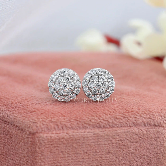 Cluster Round Cut Diamond Stud Earrings displayed on a soft pink fabric surface with a delicate background of blurred white and red flowers.