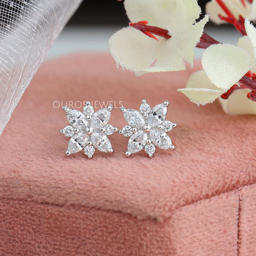 Marquise Cut Cluster Stud Earrings displayed on a soft pink cushion, featuring intricate floral designs with sparkling lab-grown diamonds, and delicate white flowers in the background.