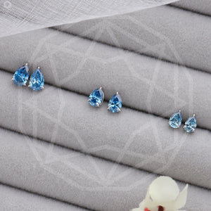 Blue Pear Shaped Diamond Stud Earrings in three sizes, arranged on a soft, gray fabric with a delicate floral accent in the corner.